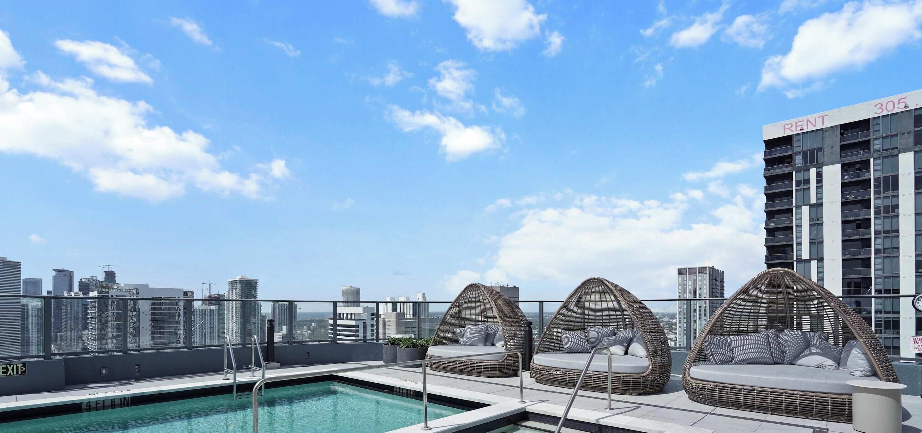 A rooftop pool with clear blue water, surrounded by white tiled flooring and a glass barrier. There are three wicker lounge chairs with cushions.