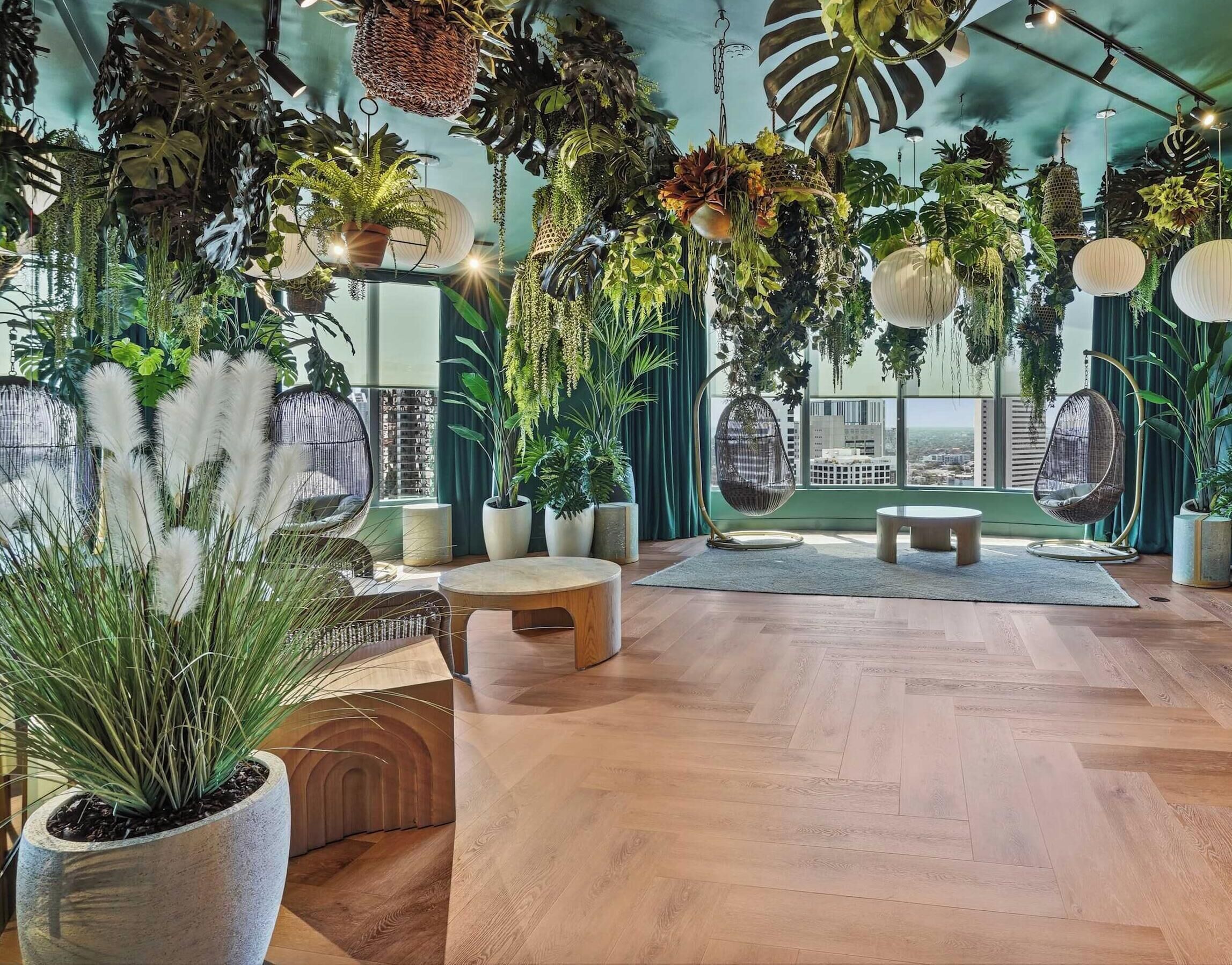A room with a herringbone wooden floor, hanging plants, a wicker chair, and a view of the city through large windows.