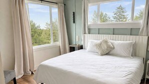 Ein Schlafzimmer mit einem Bett, zwei Fenstern, einem Nachttisch und Blick auf Bäume.