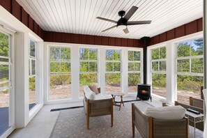 A room with large windows, wicker furniture, a ceiling fan, and a wood-burning stove. - The Cherry Cottages (Fish Creek)