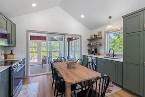 A kitchen with a dining table, chairs, and a view through glass doors. - The Cherry Cottages (Fish Creek)