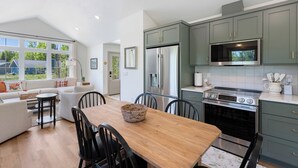 A modern kitchen with stainless steel appliances, a dining area with a wooden table, and light-colored cabinetry. - The Cherry Cottages (Fish Creek)