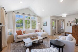 A living room with a sofa, coffee table, and chairs, a large window with curtains, and a door leading to another room. - The Cherry Cottages (Fish Creek)