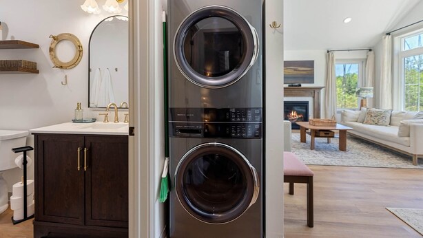 A modern laundry room with a stacked washer and dryer, a vanity with a mirror, and a fireplace in the living area. - The Cherry Cottages (Fish Creek)