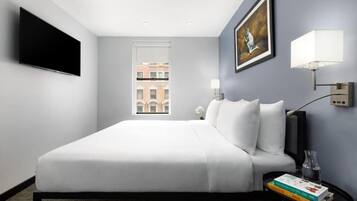 Habitación de hotel con cama, televisión de pantalla plana, un cuadro, una ventana y una mesita de noche con libros y un florero.