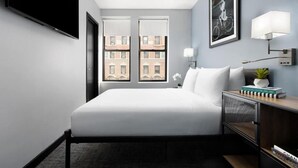A modern hotel room with a large bed, a flat-screen TV, a desk with a lamp, and a view of buildings through the windows.