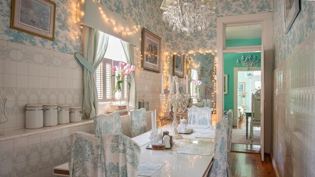 Uma sala de jantar com uma mesa posta para quatro pessoas, cadeiras com estofado floral, um lustre e luzes de cordão.