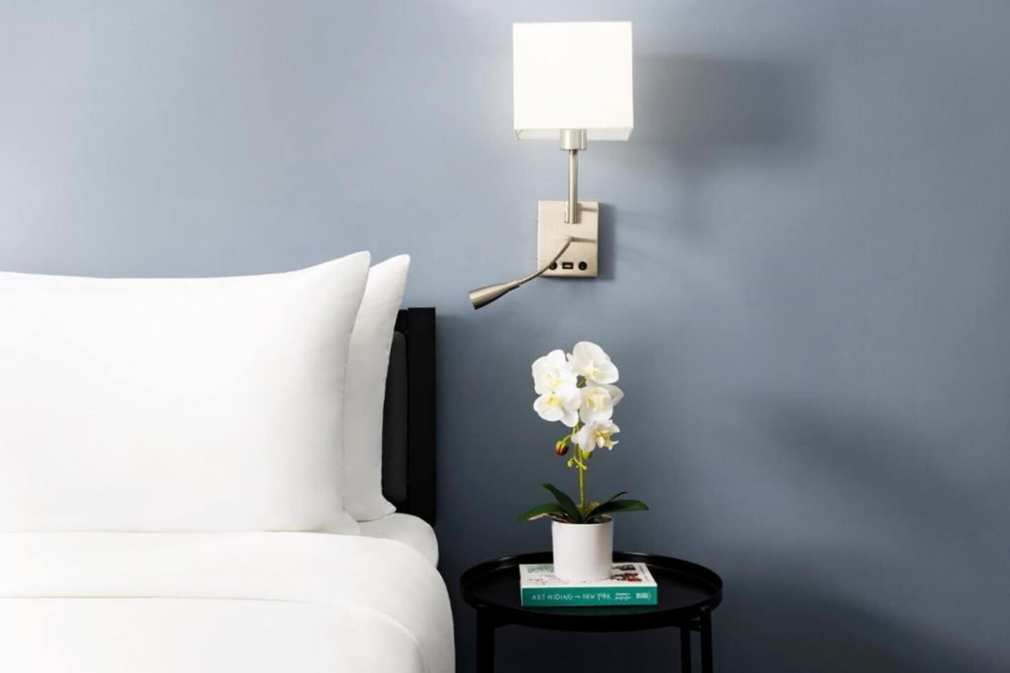 Een bed met witte beddengoed, een klein zwart nachtkastje met een witte bloevas en een boek, en een wandlamp.