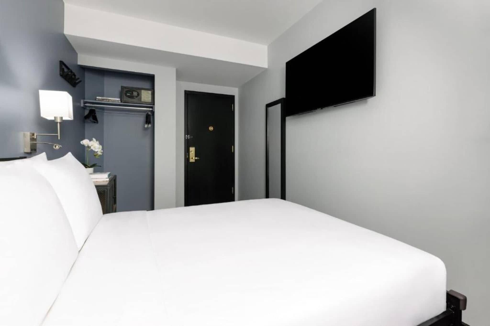 Een hotelkamer met een groot bed, een flatscreen televisie aan de muur, een klein nachtkastje met een lamp, en een deur naar een andere kamer.