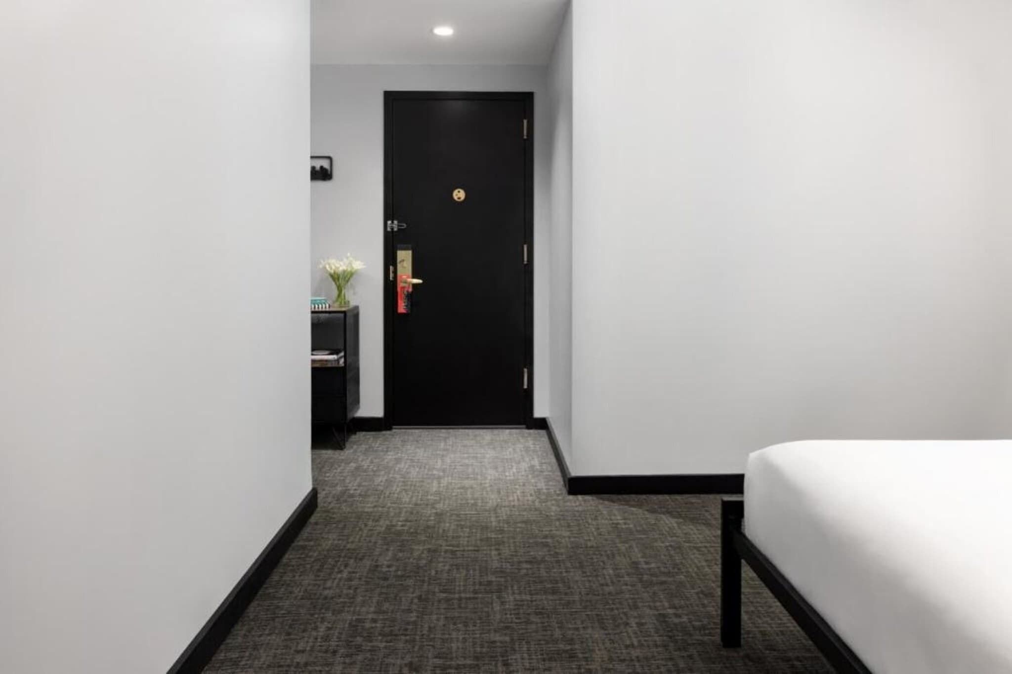 A hotel room with a bed, a desk with a vase of flowers, and a door with a lock.