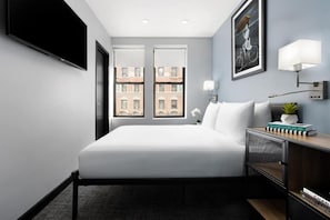 A modern hotel room with a large bed, a flat-screen TV, a desk with a lamp, and a view of buildings through the windows. - Smart Stay | Full Bed| Time Square (New York)