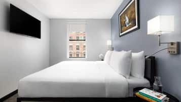 Habitación de hotel con cama, televisión de pantalla plana, un cuadro, una ventana y una mesita de noche con libros y un florero.