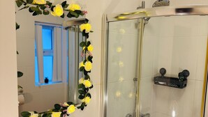 Et badeværelse med en glasafskærmet brusekabine, en vask med hane, et spejl med blomsterudsmykning og et vindue med gardiner.