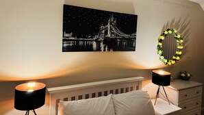 Et soveværelse med en seng, to sengeborde, en lampe, et billede i ramme af en bro og en krans på væggen.