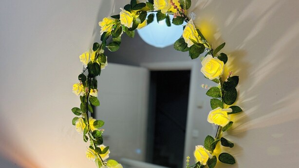 Et spejl med en blomsterkrans, en tændt lampe og en lille vase med hvide blomster på en hvid kommode.
