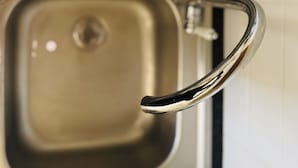 En rustfritslået vask med en kromarmatur, en gitterhylde over og en hvid flisevæg i baggrunden.