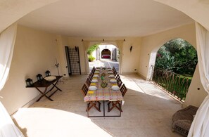 Ein Essbereich mit einem langen Tisch, gedeckt für mehrere Personen, einem kleinen Tisch mit einigen Gegenständen und Blick auf einen Garten durch einen Bogen.