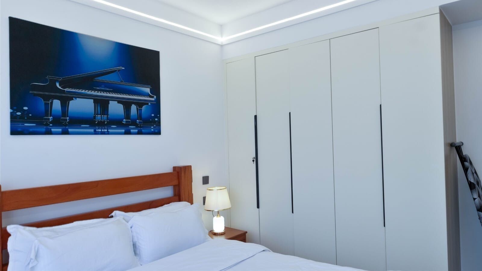 Een slaapkamer met een bed, nachtkastje, lamp, kledingkast en een schilderij van een piano.