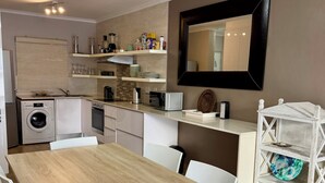 Cocina compacta con lavadora, microondas y cafetera. Hay una mesa de comedor con sillas y un armario blanco con estantes.