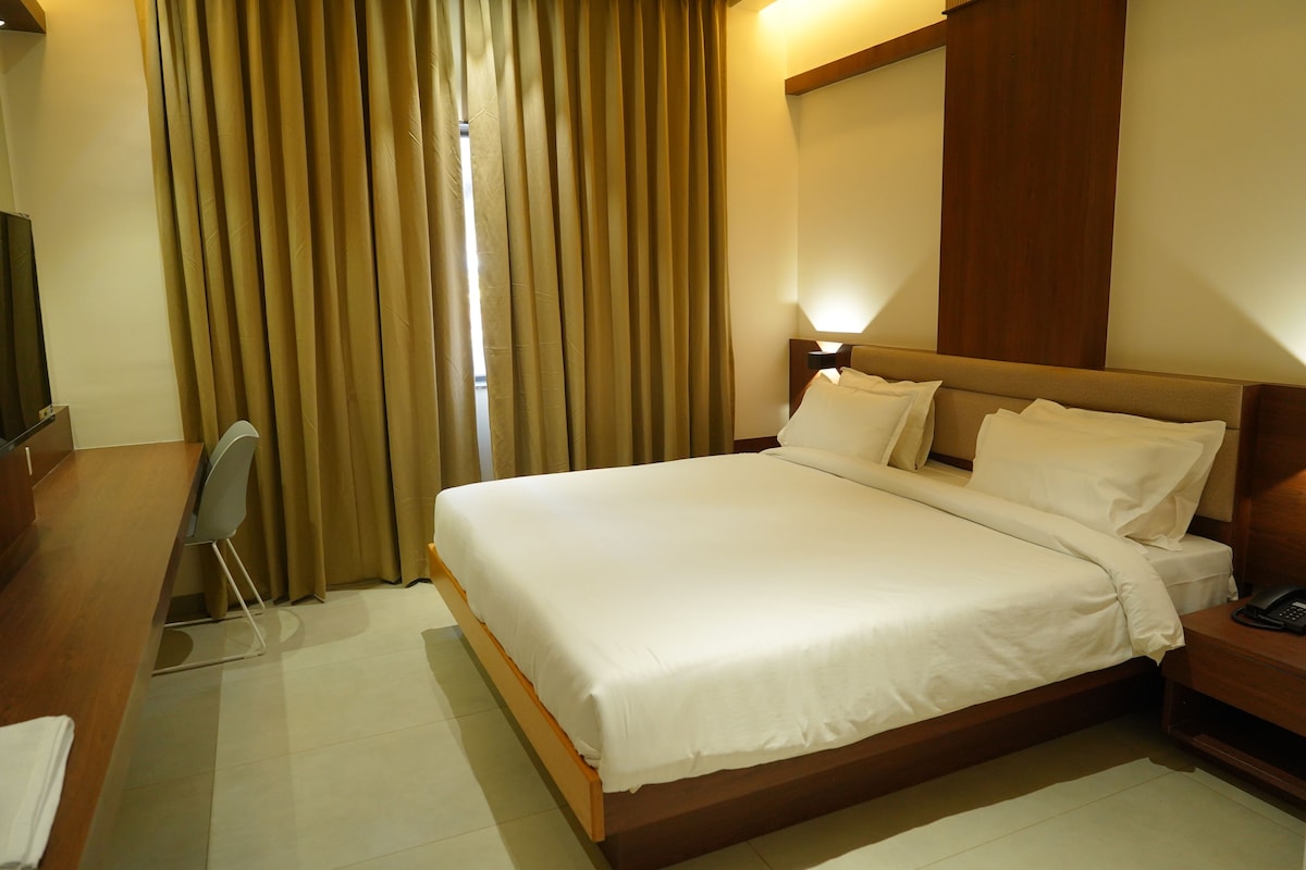 A hotel room with a large bed, a bedside table with a phone, a desk with a chair, and a TV.