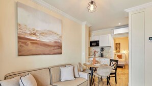 Una sala de estar moderna con un sofá color beige, una mesa de comedor con sillas, una zona de cocina con armarios blancos y un cuadro abstracto grande en la pared.