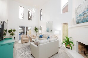 Een ruime woonkamer met een witte bank, een beige fauteuil en een open haard.