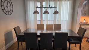 Une salle à manger avec une table rectangulaire, six chaises, une horloge murale et une cuisine visible à travers une fenêtre ornée de rideaux légers.