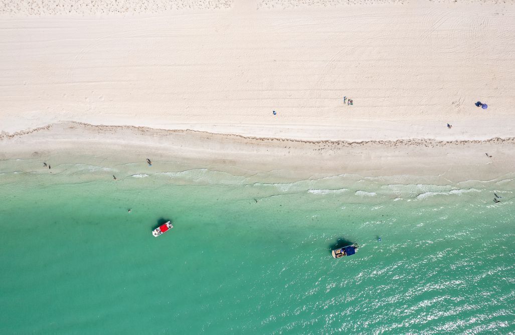 Vista aérea de una playa con agua turquesa cristalina, una orilla arenosa y algunas personas y embarcaciones.