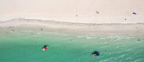 Vista aérea de una playa con agua turquesa cristalina, una orilla arenosa y algunas personas y embarcaciones.