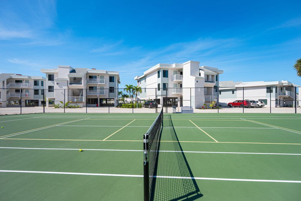 A tennis court with a net in the foreground, surrounded by multi-story buildings with balconies.