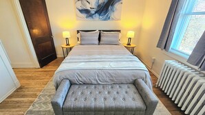 Un dormitorio con una cama, dos mesitas de noche, un banco, un radiador y un cuadro abstracto grande en la pared.