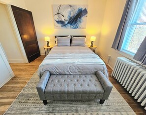 A bedroom with a bed, two bedside tables, a bench, a radiator, and a large abstract painting on the wall.