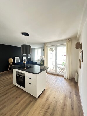 A modern kitchen with a central island, a dining area with a blue sofa, and a view of the outside through large windows. - Bright new T2 apartment next to the beach (Nice)