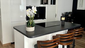 A modern kitchen with white cabinetry, a black countertop, a central island with bar stools, and a stainless steel range hood. - Bright new T2 apartment next to the beach (Nice)