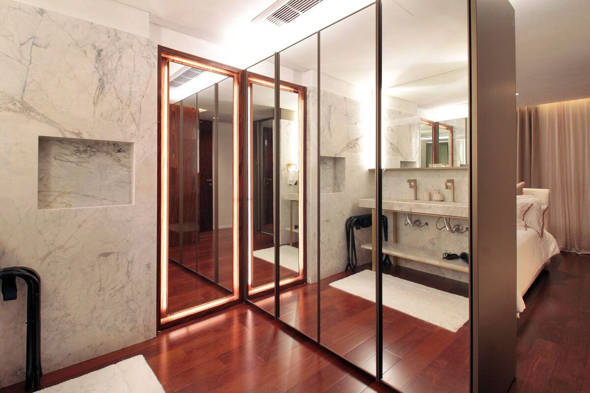 A modern hotel room with a large mirror, wooden flooring, marble walls, and a separate bathroom area.