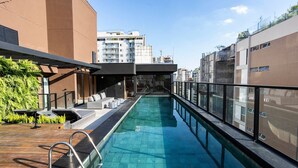 Una piscina sul tetto con terrazza in legno, ringhiere in vetro e vista sugli edifici circostanti.