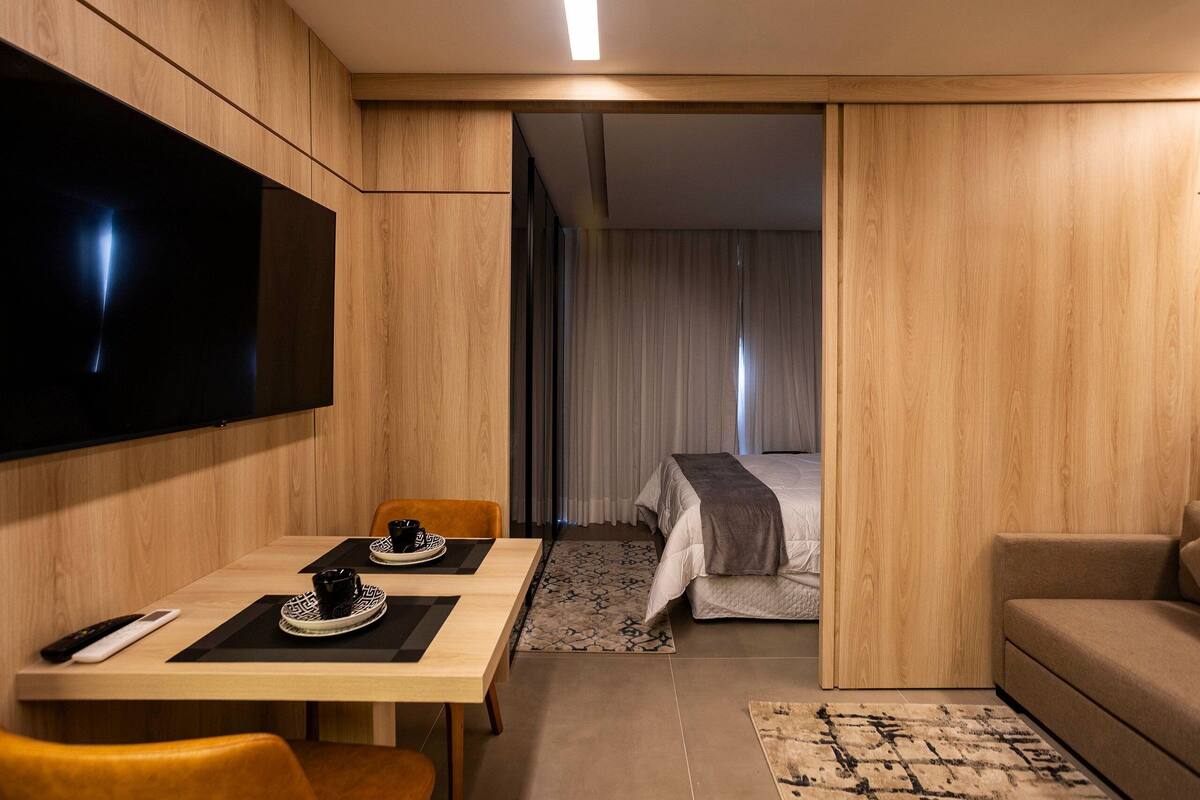 A modern hotel room with a wooden floor, a flat-screen TV mounted on the wall, a bed with a gray blanket, a sofa, and a dining table with chairs.