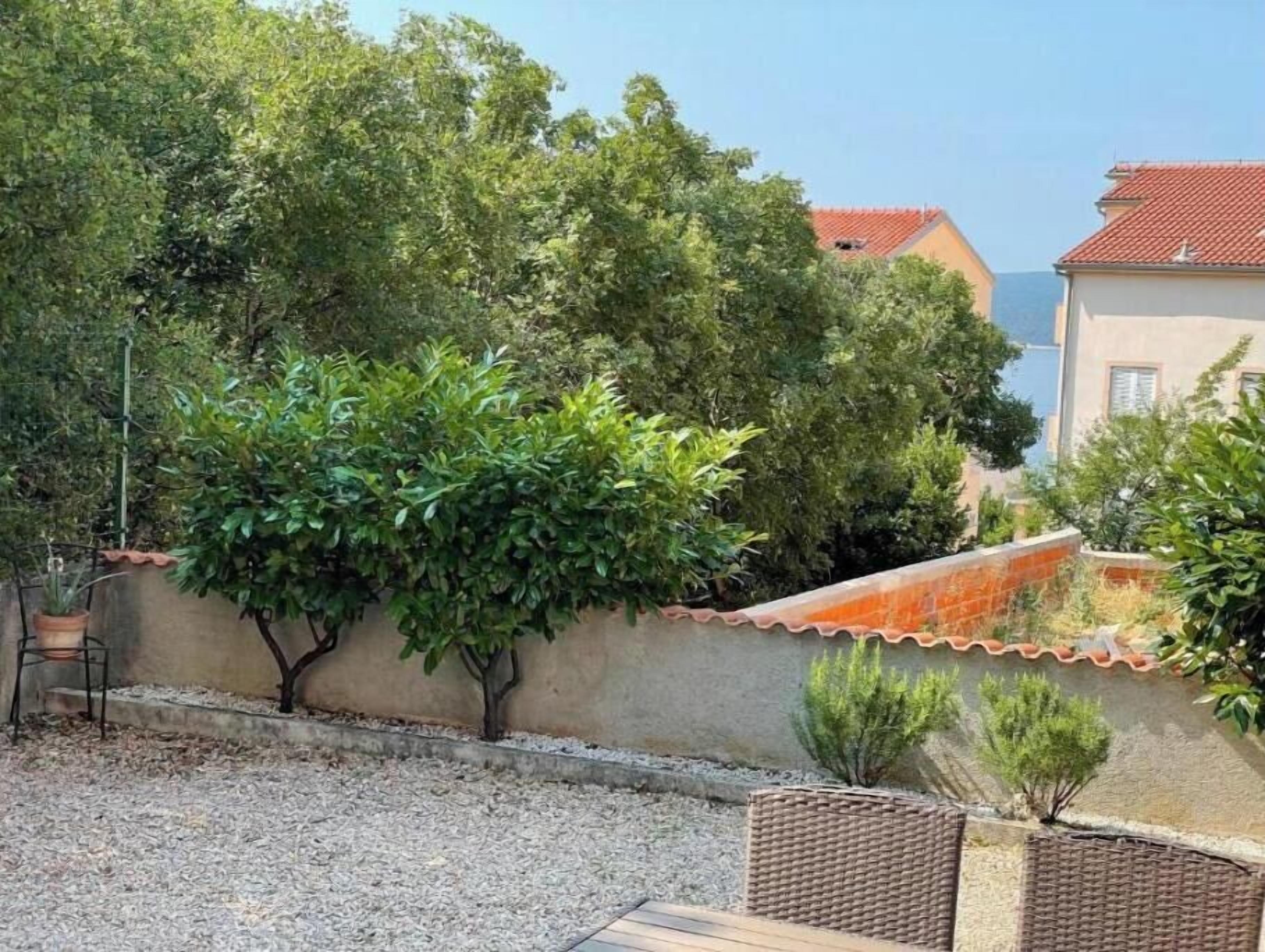 Une cour avec un sol de gravier, un petit muret en briques et différentes espèces d’arbres.