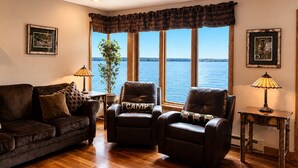 Un salon avec un plancher en bois, deux fauteuils en cuir, un canapé marron, une plante en pot et une vue sur l’eau.