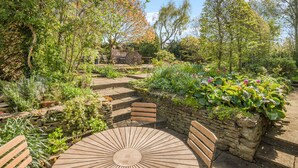 Un jardín con una mesa de madera redonda y bancos, rodeado de muros de piedra y diversas plantas.