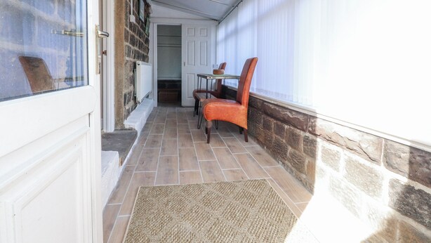 A narrow corridor with a stone wall, a door, a small table with chairs, and a window with blinds. - Vasey Croft (Keighley)