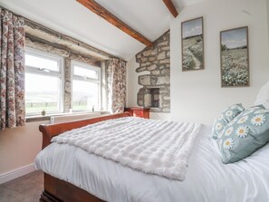 A bedroom with a stone wall, a bed with white bedding, floral curtains, and two framed pictures on the wall. - Vasey Croft (Keighley)