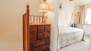 A bedroom with a wooden dresser, a bed with a white blanket, a lamp, and a window with curtains. - Vasey Croft (Keighley)