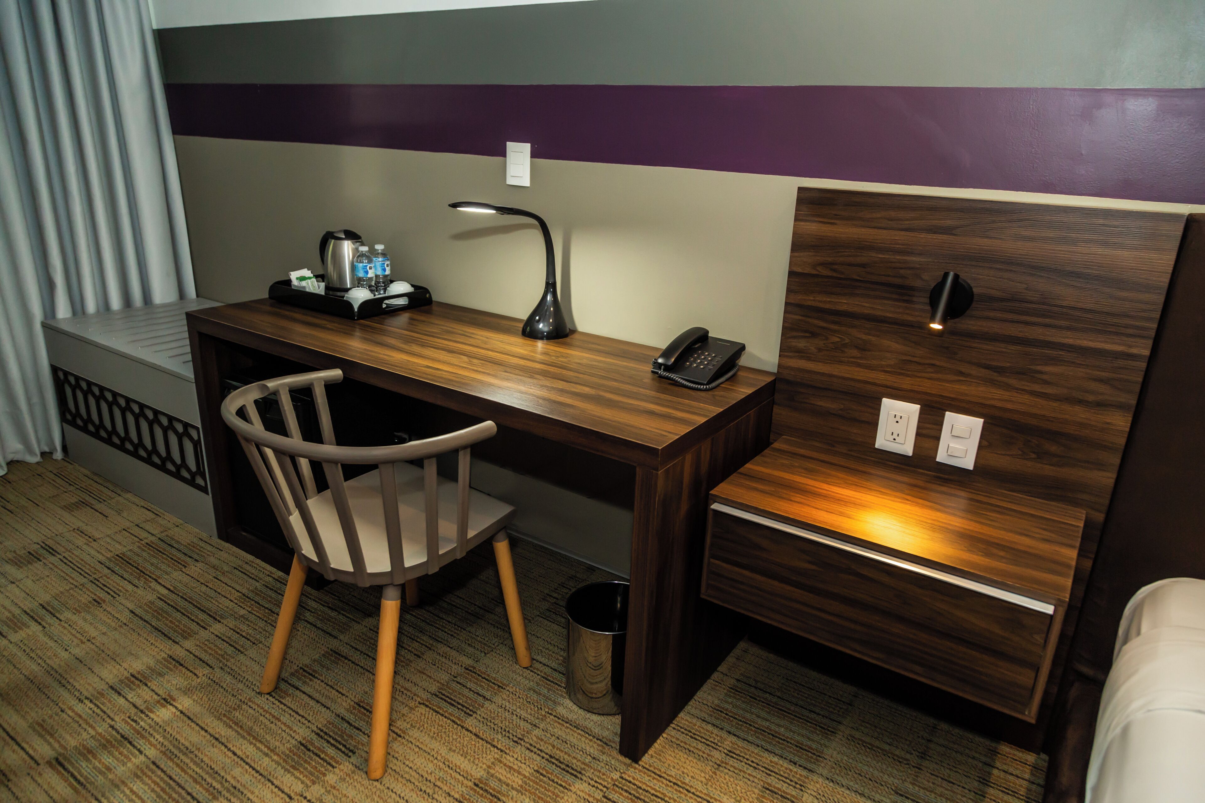 A hotel room with a wooden desk, a chair, a desk lamp, a telephone, and a small trash can.