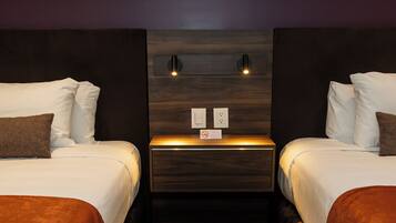 Habitación de hotel con dos camas, cada una con ropa blanca y almohadas marrones, un cabecero de madera y una mesita de noche con una lámpara.