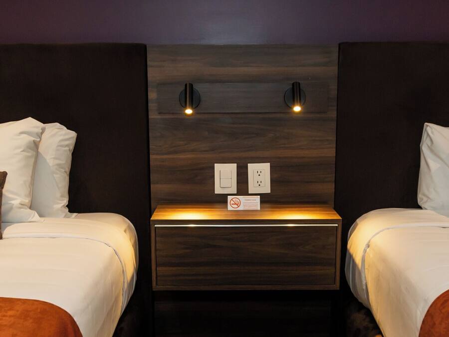Quarto de hotel com duas camas, cada uma com roupa de cama branca e almofadas castanhas, uma cabeceira de madeira e uma mesa de cabeceira com luz.
