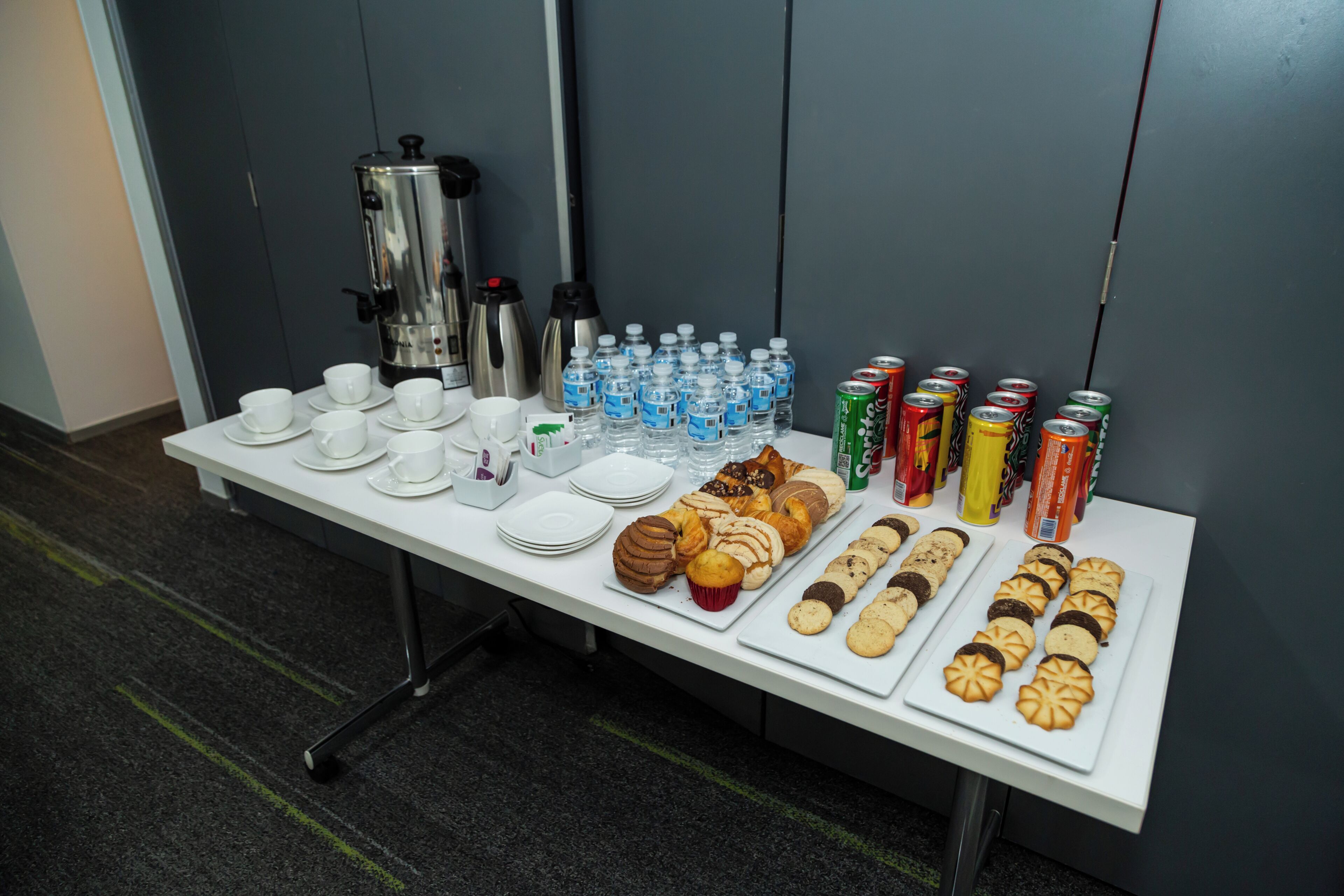 Una mesa de buffet con cafeteras, botellas de agua y diversos aperitivos.