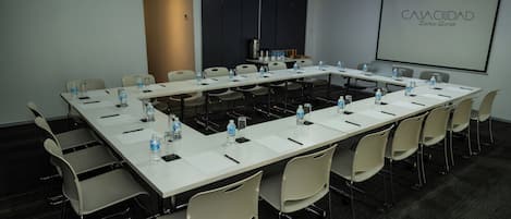 Un salón de conferencias con una larga mesa, sillas, botellas de agua y una pantalla de proyector que muestra 'CASA CIUDAD Zona Rosa'.
