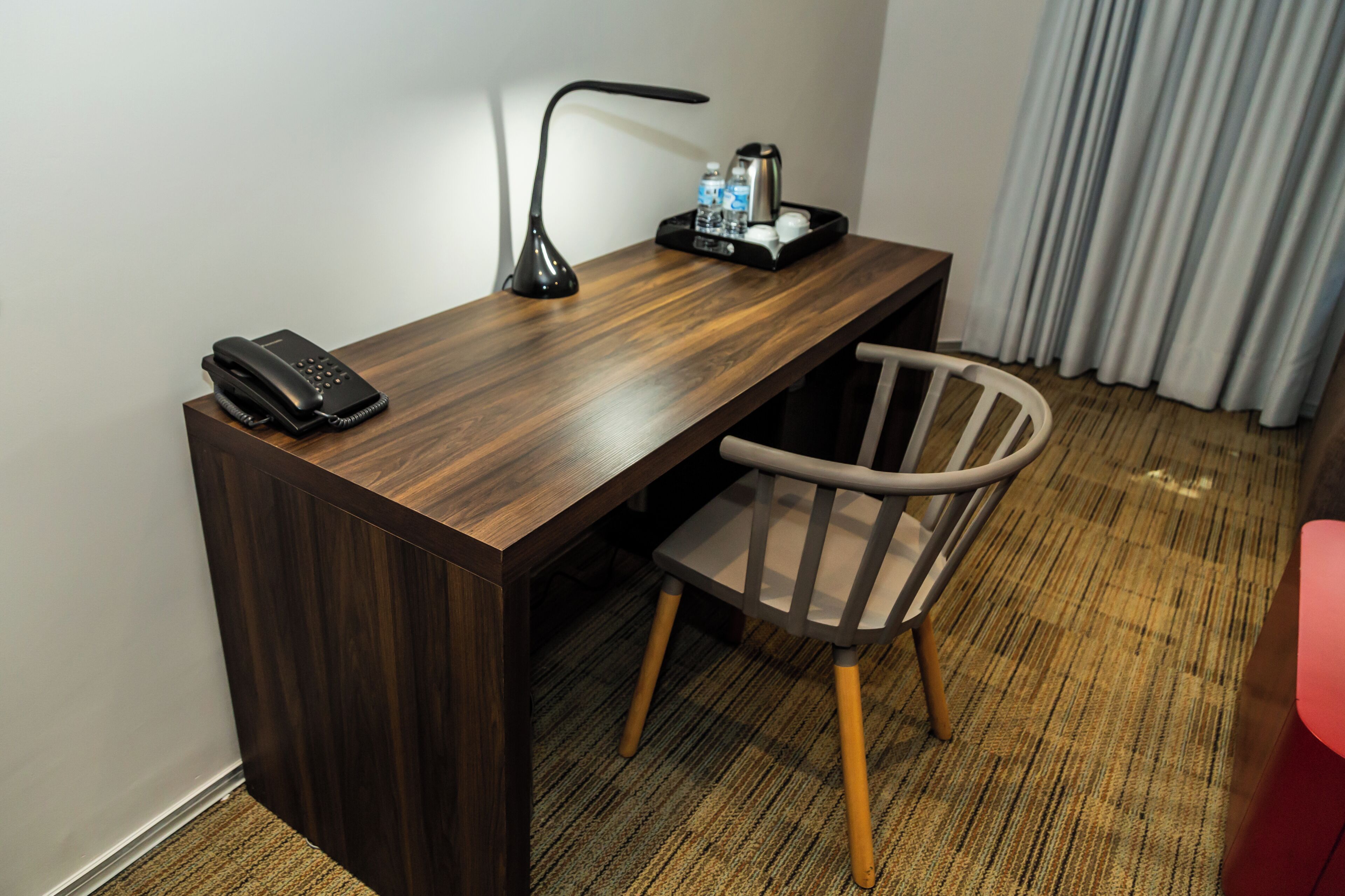 A hotel room with a wooden desk, a telephone, a lamp, a chair, and a tray with a coffee maker and water bottles.