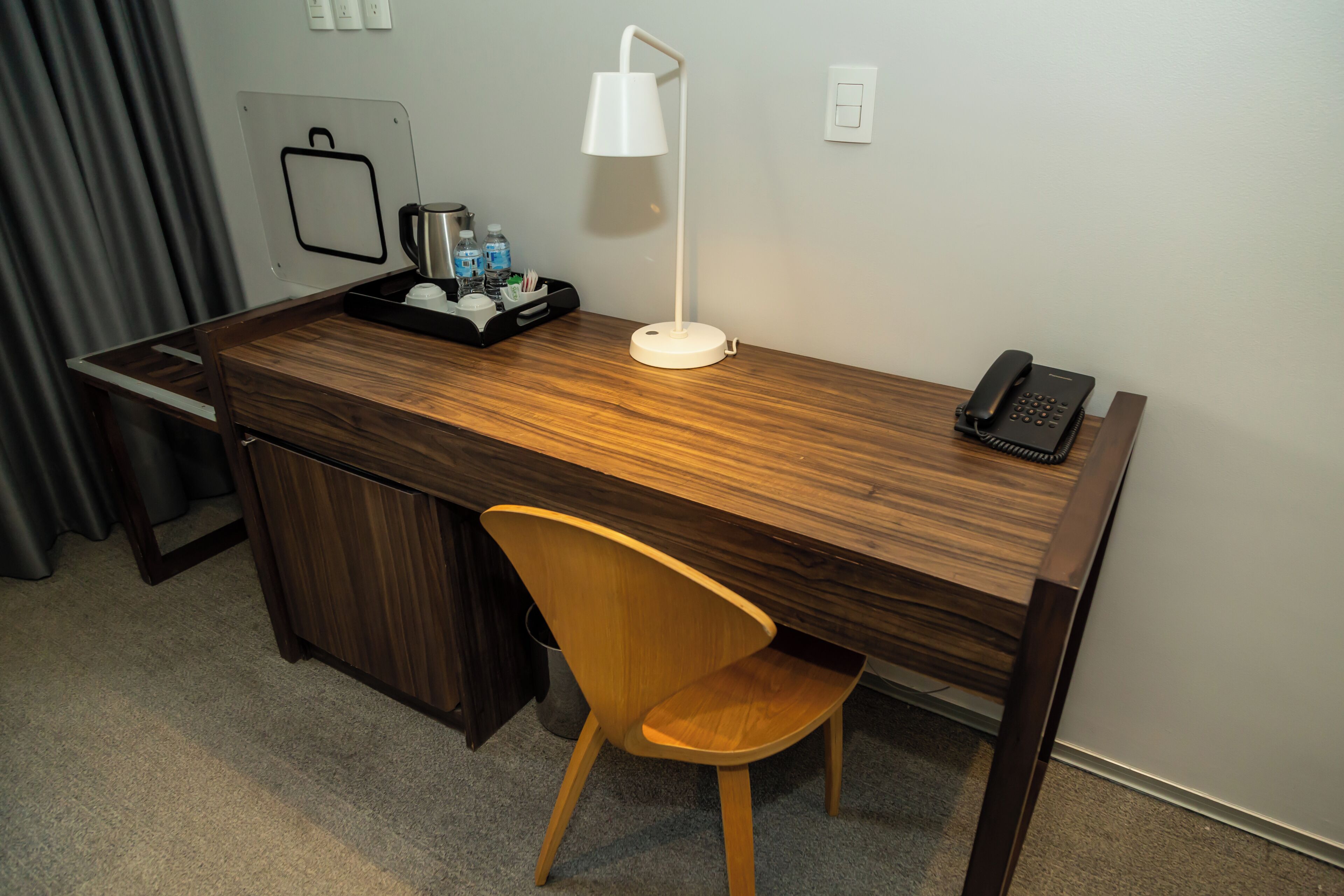 A hotel room with a wooden desk, a white desk lamp, a telephone, a chair, a tray with toiletries, and a wall-mounted power outlet.